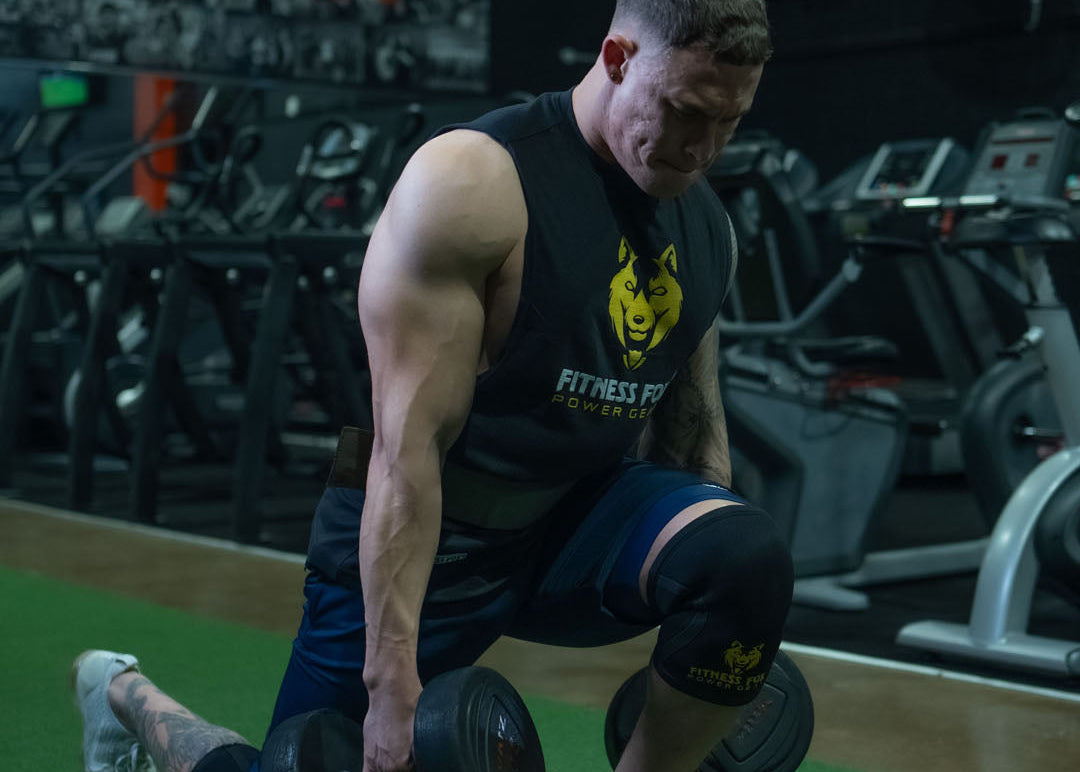 Man lifting weights in a gym wearing a 'Fitness First' tank top.